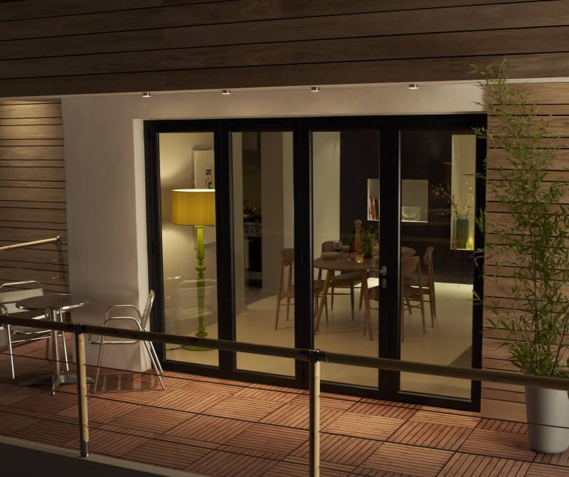 View of an elegantly furnished modern dining room through large Origin bi-fold doors that are partially open, connecting an outdoor balcony with wooden decking to the indoor space. The interior is warmly lit, featuring a dining table set for dinner and a stylish yellow floor lamp, enhancing the cosy atmosphere.